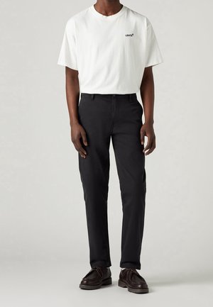 Personne portant un t-shirt blanc Levi's, un pantalon noir et des chaussures à lacets brun foncé sur un fond clair et uni.