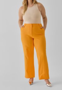 Pantaloni arancioni su misura con tasche e design a gamba dritta, abbinati a un top beige senza maniche e tacchi metallici a punta. Tessuto dalla texture liscia.