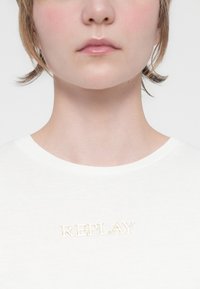 Valkoinen puuvillapaita, jossa on pyöreä kaula-aukko ja rinnan kohdalla vaaleanbeige brodeerattu "REPLAY"-logo. Yksinkertainen ja sileä rakenne.