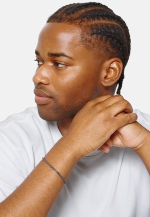 Bracelet noir en tresse au poignet, design en chaîne argentée. Sujet portant une chemise blanche, avec des cheveux tressés et un fond neutre.