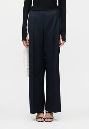 Sorte bukser med vide ben og forreste presning, båret med sort langærmet top og sorte åbne sandaler, hvidt blonderstof i siden.