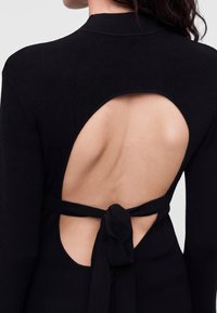 Femme portant un haut noir à manches longues avec une grande découpe ovale dans le dos, nouée à la taille avec un nœud en tissu noir.