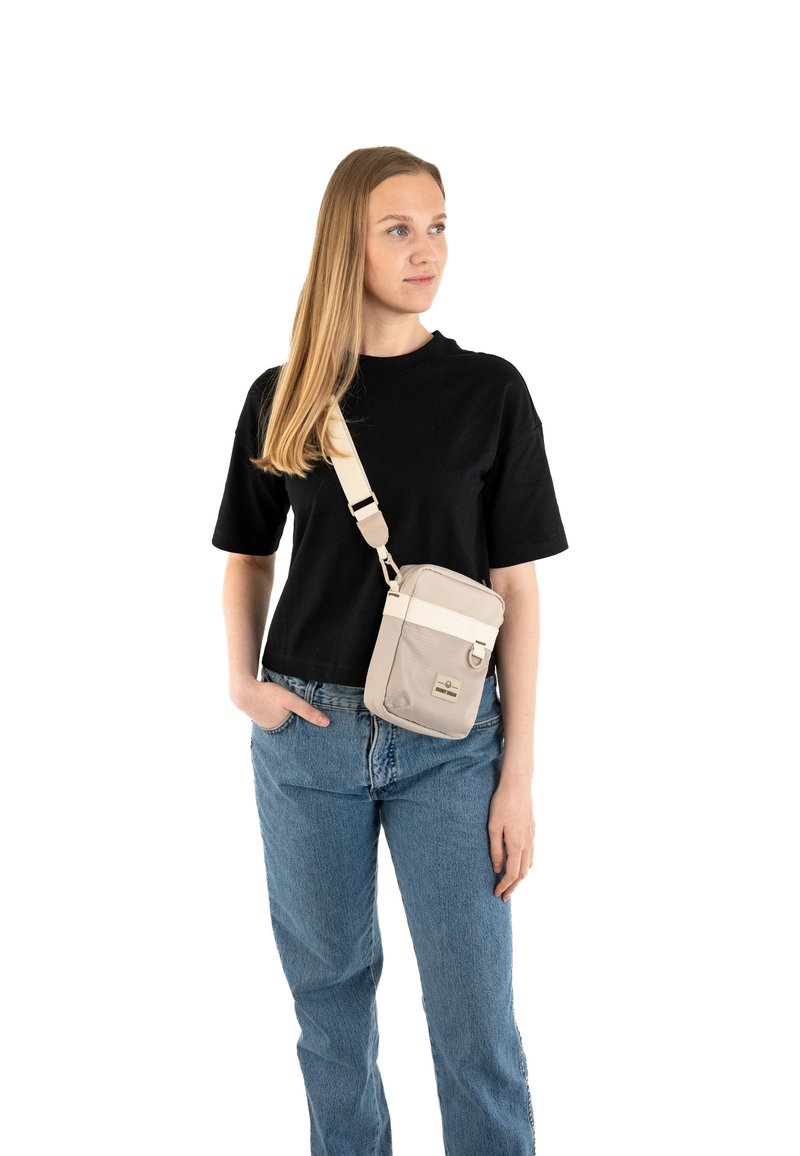 Borsa a tracolla in tessuto beige chiaro con tasca frontale, tracolla regolabile e patch con logo. Il modello indossa una maglietta nera e jeans blu.