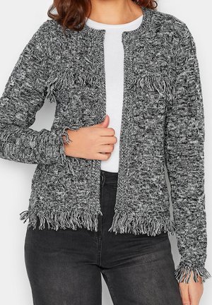 Cardigan en tricot à carreaux noirs et blancs avec des franges aux poignets, à la poitrine et à l'ourlet, porté par-dessus un haut blanc à col rond et un pantalon noir.