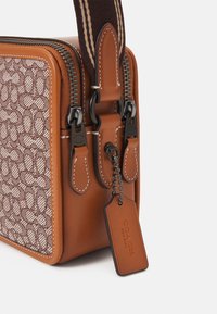 Brązowa torba crossbody z tekstylnego materiału z skórzanymi akcentami, z wzorzystą powierzchnią, podwójnymi suwakami i metką z wytłoczonym logo. Pasek w paski.