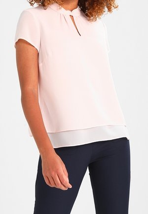 Femme portant un chemisier rose pâle à manches courtes avec un col en forme de goutte d'eau sur un pantalon bleu marine foncé, debout devant un fond blanc.
