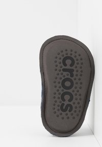 Semelle d'une chaussure Crocs gris foncé avec des points de traction circulaires et le logo "Crocs", photographiée sur un fond blanc à coins.