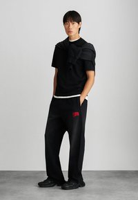 Czarna koszulka z krótkim rękawem, biała lamówka, czarne spodnie z czerwonym logo, sweter z materiału zarzucony na ramionach, czarne sneakersy. Gładki, swobodny wygląd.