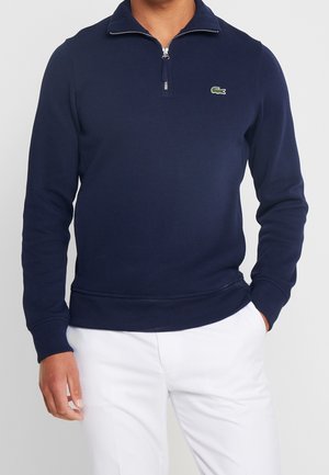 Man draagt een marineblauwe pullover met een kwart rits, voorzien van een klein groen krokodillenlogo, en witte broek, met zijn hand in zijn zak tegen een effen achtergrond.