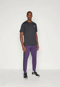 Juoda sportinė marškinėliai su mažuoju baltu logotipu, derinami su purpurinėmis sportinėmis kelnėmis, turinčiomis juodas šonines juosteles, ir juodais sportiniais bateliais.