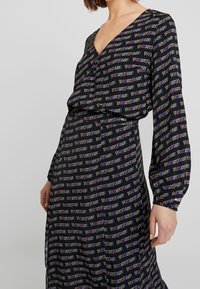 Žena v černých dlouhých šatech s vzorem tvořeným malým barevným textem "TOMMY JEANS" opakovaným po celé látce.