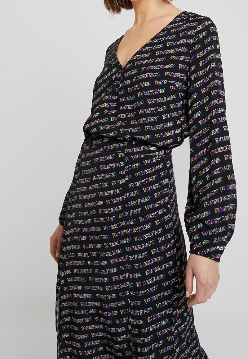 Žena v černých dlouhých šatech s vzorem tvořeným malým barevným textem "TOMMY JEANS" opakovaným po celé látce.