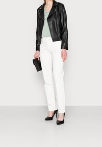 Schwarze Leder-Motorradjacke, hellgrünes V-Ausschnitt-Shirt, weiße gerade Jeans und schwarze spitze Pumps. Schwarze Clutch in der Hand.