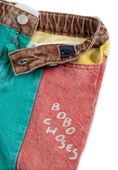 Pantalons en denim multicolores avec des sections teal, rouge et jaune, avec des coutures brunes et "BOBO CHOSES" imprimé en gris sur le tissu rouge.
