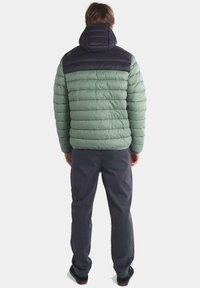 Chaqueta acolchada verde con capucha y sección de hombros en azul marino, presenta un diseño acolchado y un tejido texturizado. Se lleva con pantalones gris oscuro.