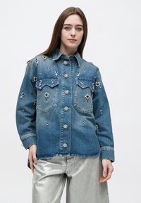 Νεαρή γυναίκα με μπλε τζιν μπουφάν διακοσμημένο με πέρλες και κοσμήματα, συνδυασμένο με λαμπερό ασημί παντελόνι, σε απλό φόντο.