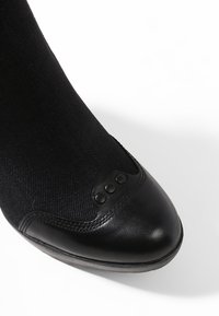 Zapato negro con punta de cuero liso y parte superior de tela negra texturizada, con tres acentos redondeados cerca de la zona de los dedos. Diseño de perfil bajo.