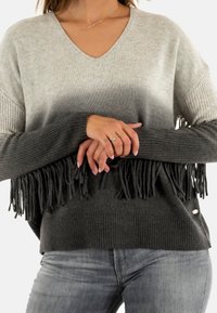 Maglione grigio ombre con scollo a V, maniche a costine e orlo frangiato. Tessuto morbido e vestibilità rilassata, completato da un discreto dettaglio di branding.