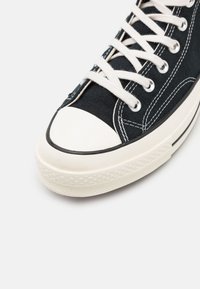 Sneaker alta in tela nera con punta in gomma bianca, lacci bianchi e cuciture dettagliate. Suola piatta con grip testurizzato.