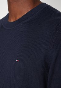 Gros plan d'une personne portant un pull en tricot bleu marine avec un petit logo rectangulaire rouge, blanc et bleu sur la poitrine.