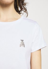 Žena v bílém tričku s designem hmyzu z kamínků na levé horní části hrudi a se zlatým visacím náušnicí na levém uchu.