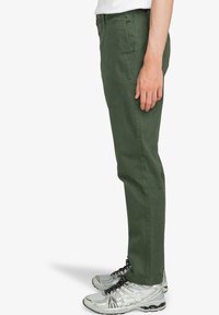 Pantaloni in cotone verde con design a gamba dritta, dotati di tasche laterali e una texture liscia, abbinati a scarpe sportive grigie.