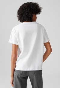 Weißes Baumwoll-T-Shirt mit umgeschlagenen kurzen Ärmeln, rundem Ausschnitt und lockerer Passform. Die Rückansicht zeigt keine zusätzlichen Muster oder Akzente.