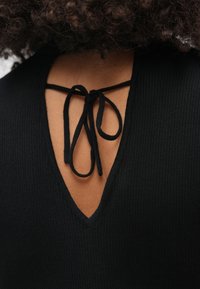 Närbild av ett svart ribbat plagg med en djup V-ringning och ett knutet svart snöre baktill, båda bärs av en person med lockigt hår.