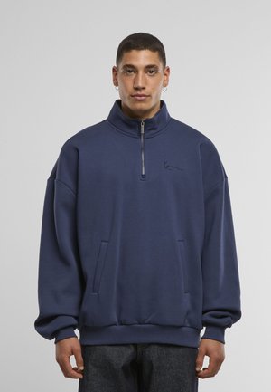 Tumesinine pullover-treeningdress koos kõrge krae, pooleldi tõmblukuga ja ees taskutega. Rinnal on peen tikandiga logo.