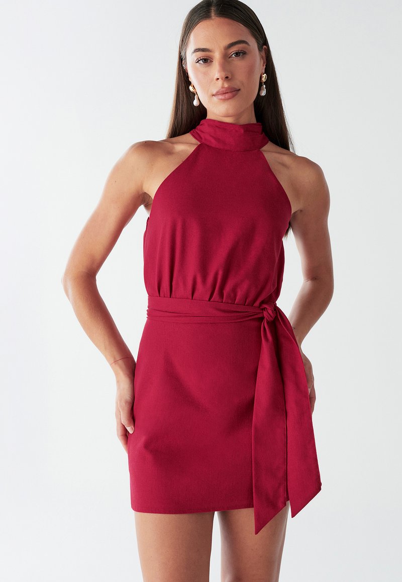 Sarkana haltera kakla kleita ar pieguļošu korseti, elastīgu vidukli un sasienamu jostu kā akcentu. Gluda auduma kleita ar vienkrāsainu rakstu un mini svārku dizainu.