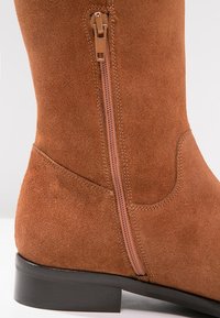 Bota alta de ante marrón, con cremallera lateral en marrón claro. Presenta un tacón bajo negro y una textura suave, enfatizando un diseño clásico.