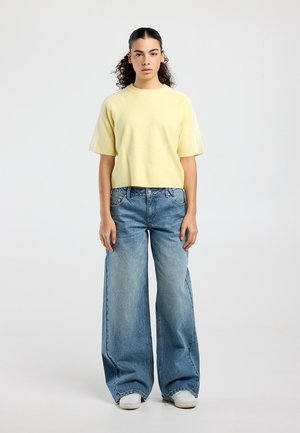 Jonge persoon staand, gekleed in een lichtgeel topje met korte mouwen, wijd uitlopende blauwe spijkerbroek en witte sneakers, tegen een effen achtergrond.