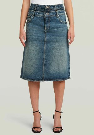 Jeans knielanger Rock mit zwei Taillenbändern und Knöpfen, kombiniert mit schwarzen Riemchenabsätzen und einem weißen Oberteil.