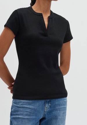 Vrouw die een zwart top met korte mouwen en een ronde hals met split draagt, en blauwe spijkerbroek, staand met de handen achter de rug.