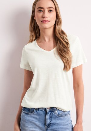 Žena s dlouhými blond vlasy, která má na sobě jednoduché bílé triko s výstřihem do V a krátkými rukávy a světlé modré džíny na světlém pozadí.