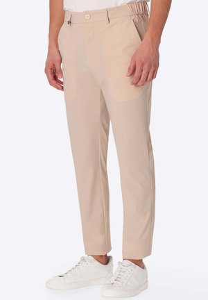 Uomo che indossa pantaloni beige slim fit con vita elastica e sneakers bianche, in piedi davanti a uno sfondo chiaro e semplice.