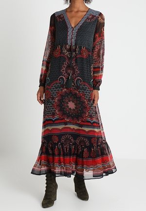 Frau trägt ein langärmliges Maxi-Kleid mit V-Ausschnitt und aufwändigen roten, schwarzen und blauen Blumen- und geometrischen Mustern, kombiniert mit olivgrünen Schnürstiefeln.