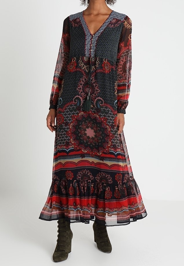Femme portant une robe longue à manches longues et col en V avec des motifs floraux et géométriques complexes en rouge, noir et bleu, associée à des bottes à lacets vert olive.