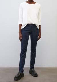 Bílá košile s tříčtvrtečními rukávy, navy modré slim-fit džíny a černé robustní polobotky. Hladká textura, minimalistický design.