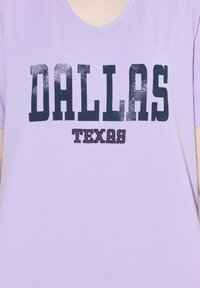 Camisa de cuello en V color lavanda con gran texto negro desgastado "DALLAS" y letras en bloque más pequeñas "TEXAS" debajo en el pecho.
