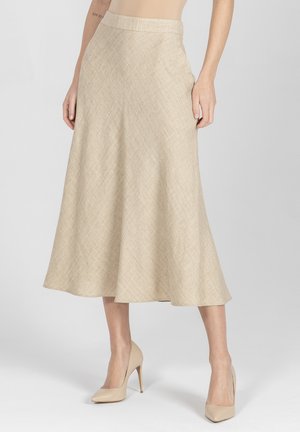 Kobieta w beżowej, rozkloszowanej spódnicy midi i beżowych szpilkach, stojąca na tle jednolitego, jasnoszarego tła.