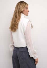 Biely bezrukávový sveter s vysokým golierom, cez priesvitnú blúzku s dlhými rukávmi, v kombinácii s námorníckymi nohavicami s bielymi bodkami a bočnými vreckami.