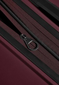 Valigia bordeaux con cerniera nera; presenta un tessuto texturizzato e un comodo tirante della cerniera, evidenziando materiali robusti e un design funzionale.