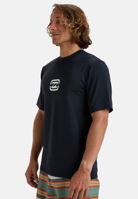 Mörkblå kortärmad rashguard med en rund halsringning och en vit logotyp i mitten. Slät, elastisk tyg; bärs med flerfärgade randiga badshorts.