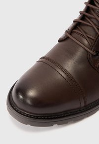 Stivale in cuoio marrone con punta arrotondata, finitura testurizzata e dettagli di cucitura; design con lacci e occhielli in metallo, suola in gomma.