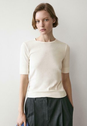 Giovane con capelli corti che indossa una maglietta crema a maniche corte e pantaloni scuri a righe sottili, in piedi contro uno sfondo chiaro e semplice.