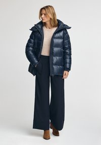 Doudoune marine à finition brillante, avec capuche et sections cousues, assortie à un top beige et un pantalon large marine. Bottines brunes.