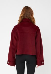 Burgundy cropped jacket made of fleece, featuring a wide collar and long sleeves with cuff accents. Smooth texture and relaxed fit.