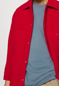 Giacca camicia rossa di lana con due tasche, chiusure a bottoni e colletto, indossata sopra una maglietta azzurra chiara.