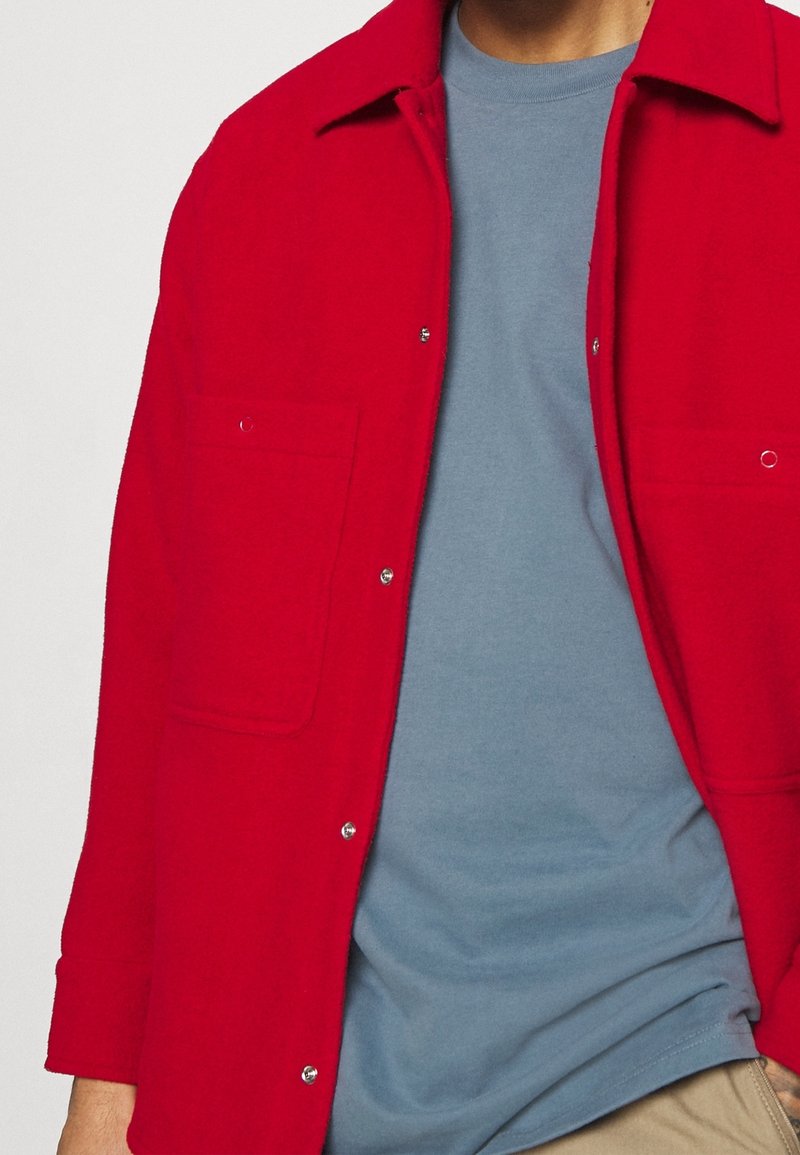 Giacca camicia rossa di lana con due tasche, chiusure a bottoni e colletto, indossata sopra una maglietta azzurra chiara.